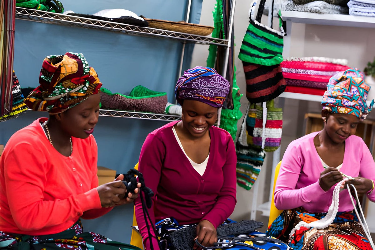 Three women wearing colorful headscarves working on knitting or similar crafts in a workshop setting.