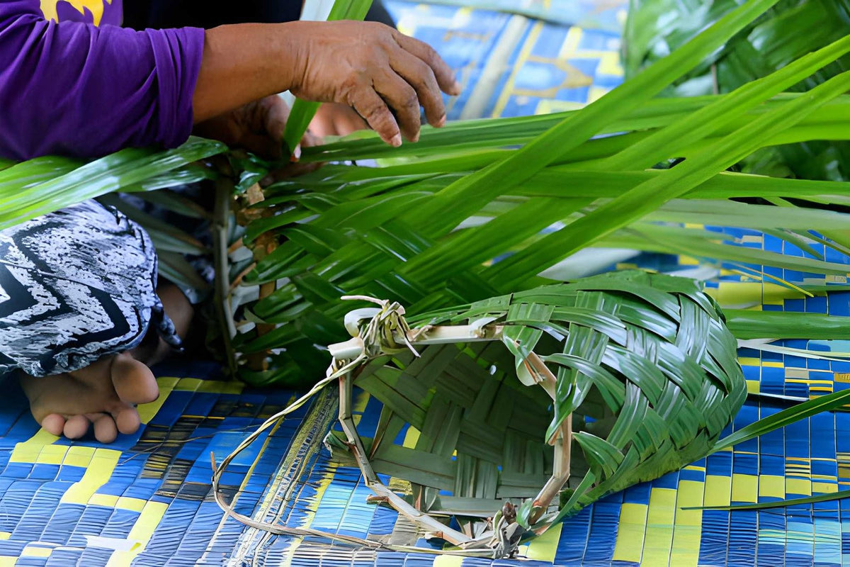 Person crafting a green woven basket using palm leaves on a blue and yellow mat.